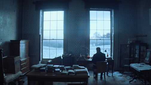 A man sits at a writing desk, facing a window that overlooks the parkland